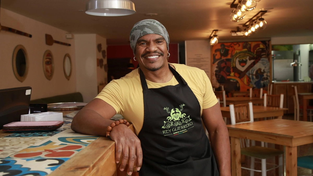 Rey Guerrero apoyado en la barra de baldosas de colores del restaurante, con el mural del Pacífico y la cocina abierta al fondo